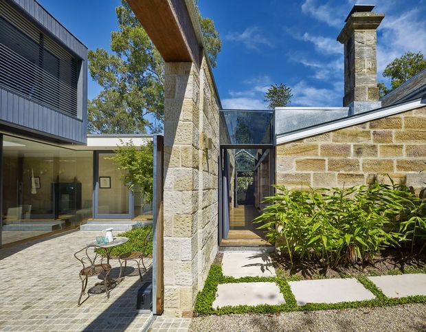 A sunny courtyard connecting a contemporary glazed addition with an older masonry structure. The scene features rough light sandstone walls, a prominent timber beam, dark metal-framed glazing, a stone-paved patio with a small outdoor table and chairs, planted garden beds, and a substantial masonry chimney. The architecture combines modern additions with retained traditional fabric.