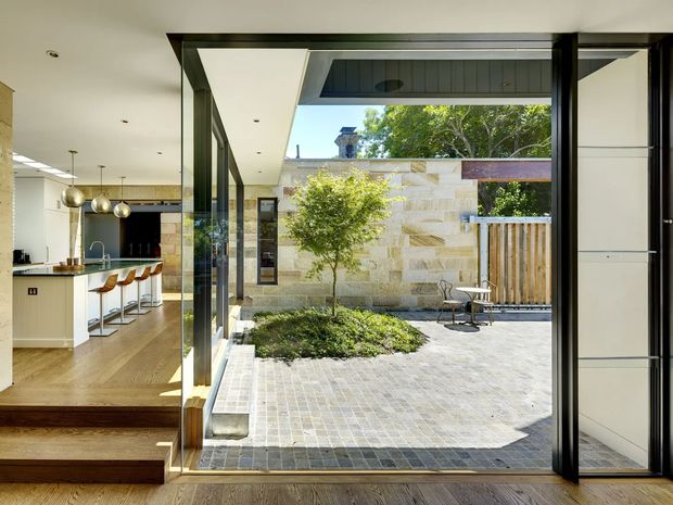 A contemporary open-plan kitchen is viewed through fully opened dark-framed glass doors onto a sheltered landscaped courtyard. The kitchen has a white island with timber stools, pale timber flooring, stainless-steel appliances and spherical pendant lights. Outside, pale stone walls, grey paving, a small planted garden bed and timber screening create a private indoor–outdoor connection.