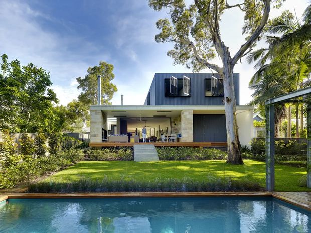 Exterior view of a contemporary two-storey residence set within a lush landscaped garden, viewed across a swimming pool. The home has a dark vertically clad upper level, pale masonry walls, broad covered outdoor living space, projecting window shutters, and a flat-roofed lower form. Mature trees and tropical planting surround the property.