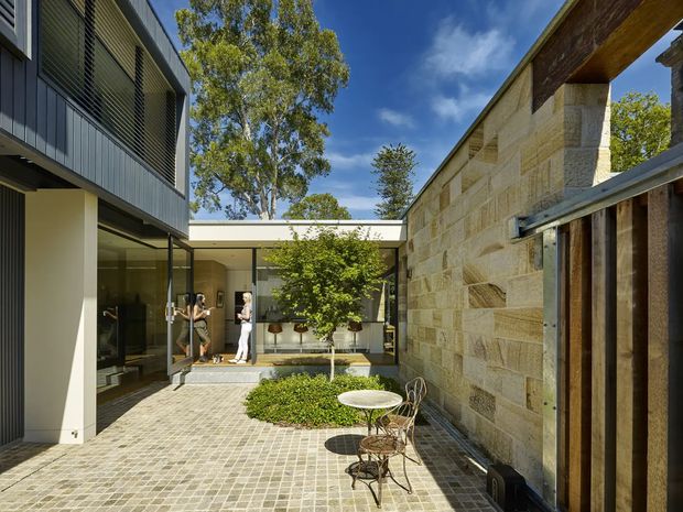 A sunlit contemporary courtyard enclosed by a modern dark-clad building and a substantial pale stone wall. The courtyard has small-format grey paving, a planted central garden bed with a young tree, and a compact outdoor café-style table and chairs. Full-height glazing opens toward an interior living or circulation space, while timber and metal detailing frame the right side. Mature trees and blue sky are visible above the courtyard walls.