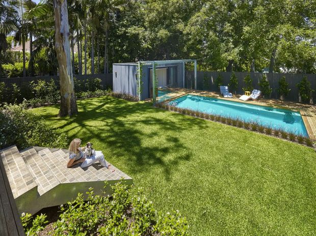 A landscaped residential backyard viewed from an elevated position, featuring a large manicured lawn, mature tropical and leafy trees, a turquoise rectangular swimming pool with timber decking, and a compact modern poolside structure with glazed openings. A paved or tiled raised terrace with seating occupies the foreground, while a dark privacy fence encloses the garden.