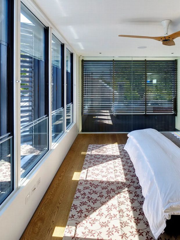 Bright contemporary bedroom with a partially visible bed, white walls and ceiling, warm timber flooring, and a patterned area rug. Full-height glazing lines one side of the room, while dark horizontal louvred blinds provide privacy and sun control. A timber-bladed ceiling fan and dark window framing add contrast, with strong sunlight creating geometric shadows across the floor and rug.