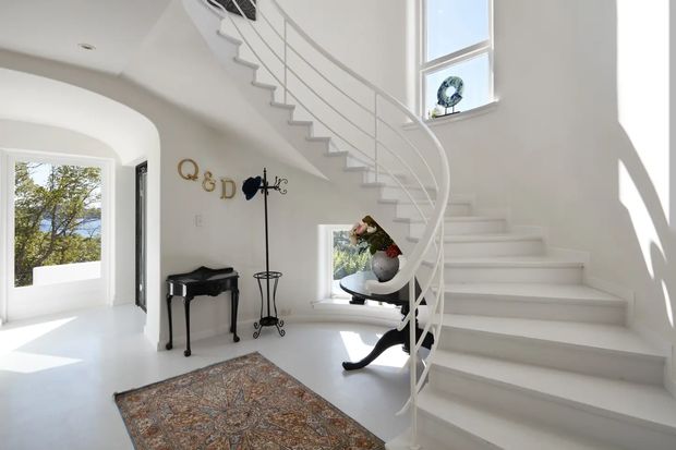 Bright, elegant residential entry hall dominated by a sweeping white curved staircase with slim metal balustrade and a tall window. The space has white walls and floors, arched openings, a dark antique-style console table, decorative wall lettering, a small side table, and a patterned area rug. Natural daylight creates a serene, refined atmosphere.