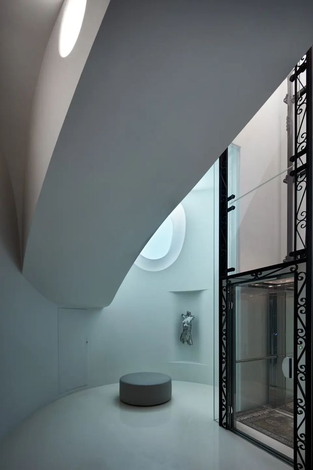 A sculptural, monochromatic interior stairway or landing with sweeping curved white forms, a smooth white floor, recessed oval skylights or light wells, a small round upholstered stool, and a decorative black wrought-iron framed glazed opening on the right. The space has a dramatic, gallery-like minimalist character.