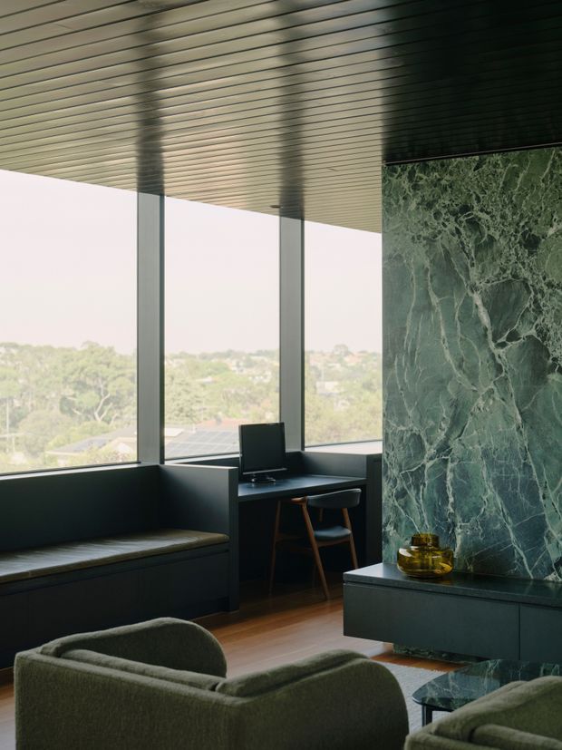 A sophisticated contemporary living room with floor-to-ceiling windows overlooking a leafy suburban landscape. Dark built-in window seating and a compact integrated desk frame the glazing, while a dramatic green veined stone fireplace wall forms the main feature. The room includes sculptural upholstered lounge chairs, a low dark hearth, timber flooring, and a linear ribbed ceiling with recessed detailing.