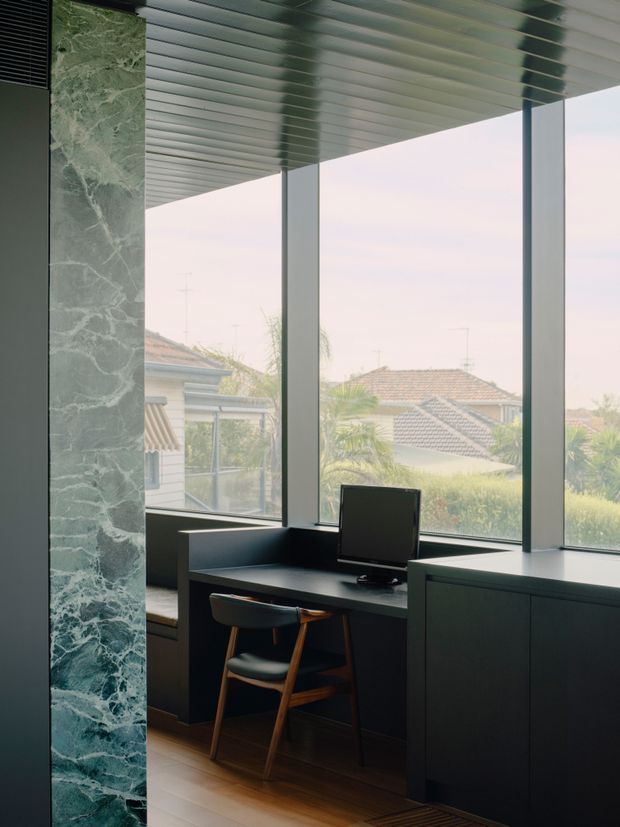 A compact home office positioned beside large floor-to-ceiling windows, with a dark built-in desk and cabinetry, computer monitor, and a simple timber-framed chair. A dramatic green veined stone-clad column frames the left side, while a ribbed metallic ceiling and slim dark window frames create a refined contemporary interior. Suburban rooftops and planting are visible outside.