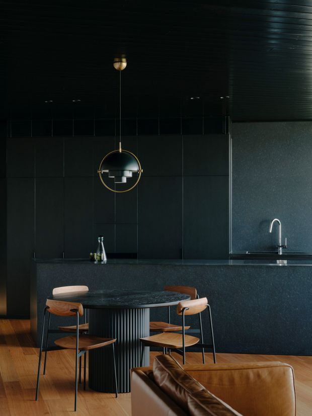 A moody, sophisticated open-plan kitchen and dining area in a dark palette. Matte black or charcoal wall and ceiling finishes surround a dark stone kitchen island with an integrated sink and curved chrome tap. A round dining table with a ribbed cylindrical base is paired with timber and black-framed chairs beneath a sculptural brass pendant light. Warm timber flooring and a tan leather sofa introduce contrast and warmth.