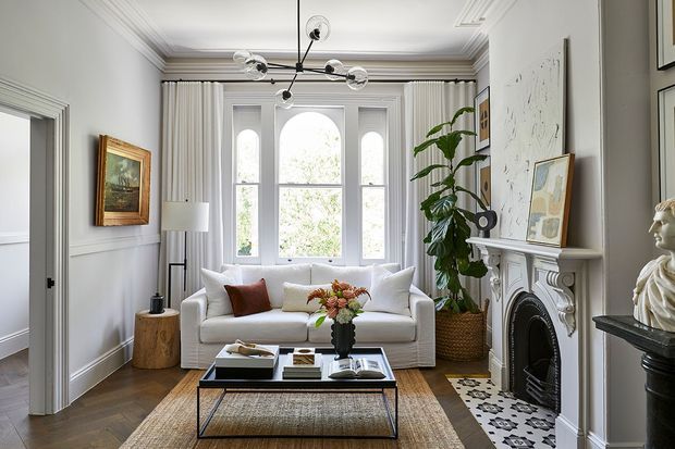 Refined traditional living room with decorative wall panelling, ornate cornices, tall curtained sash windows, a detailed white fireplace surround, and herringbone timber flooring. A contemporary sculptural chandelier, modern white sofa, black-framed coffee table, artwork, and indoor plants create a layered classic-contemporary interior.