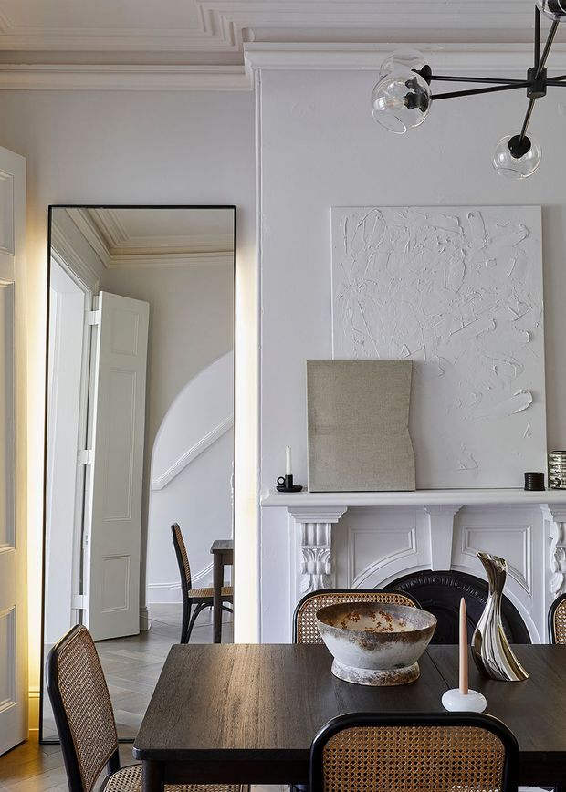 Elegant dining room in a classic apartment or townhouse, featuring a dark timber dining table with woven cane chairs, an ornate white decorative fireplace mantel, textured white artwork, and a sculptural contemporary pendant light. A large black-framed mirror reflects a paneled doorway and adjoining hall, while decorative ceiling and wall mouldings add refined character.