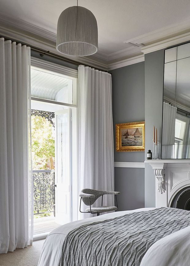 Elegant bedroom interior with a grey painted wall, ornate white fireplace mantel, mirrored overmantel, tall white curtains, detailed ceiling cornices, a sculptural pendant light, and a bed dressed in layered grey and white textiles. French doors open to a small balcony with decorative wrought-iron railing.