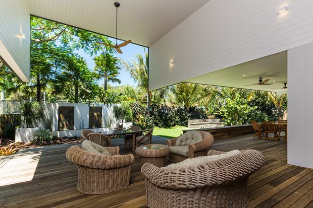 A bright, open-air covered outdoor living area with a timber deck, white-lined ceiling and walls, ceiling fans, and broad openings to a lush tropical garden. Woven rattan lounge chairs and a sofa surround low tables, while a dining setting sits further beneath the roof. The space has a relaxed resort-like character and strong indoor-outdoor connection.