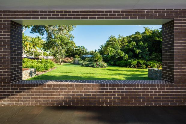 View from a covered outdoor or semi-enclosed area through a large rectangular brick-framed opening toward a landscaped garden and lawn. Dark brown brick walls, lintel, and deep sill frame the scene, with dark tiled flooring in the foreground and lush tropical planting beyond.