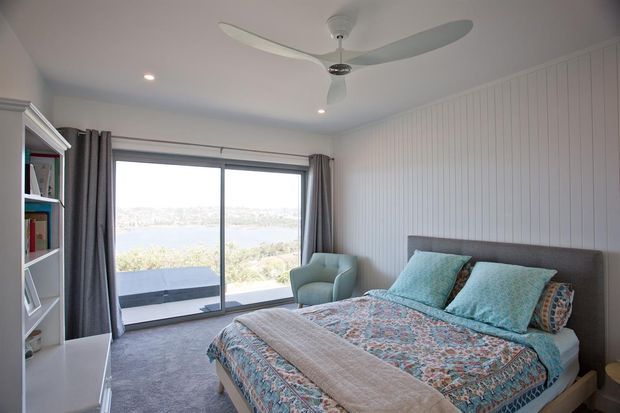 A bright, relaxed bedroom with a large sliding glass door opening to a scenic view. The room features a bed with patterned blue and coral bedding, pale blue cushions, a grey upholstered headboard, a matching armchair, built-in white shelving, carpeted flooring, white vertical wall panelling, curtains, recessed lighting, and a modern ceiling fan.