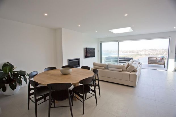 Bright open-plan interior with a round timber dining table and dark dining chairs in the foreground, flowing into a minimalist living area with a light sectional sofa and fireplace. Large sliding glazing opens to a balcony or terrace with a waterfront view, while a skylight and recessed ceiling lights provide additional daylight.
