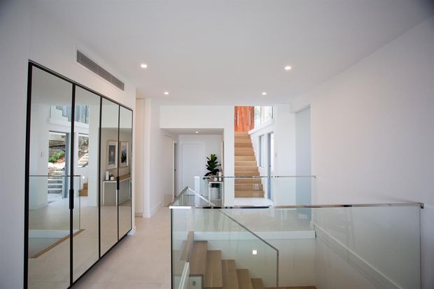 Bright contemporary interior showing a stair landing and hallway with pale flooring, white walls, a glass balustrade, timber stair treads, and a black-framed mirrored built-in wardrobe or storage wall. A double-height void and upper-level glazing bring in abundant natural light.