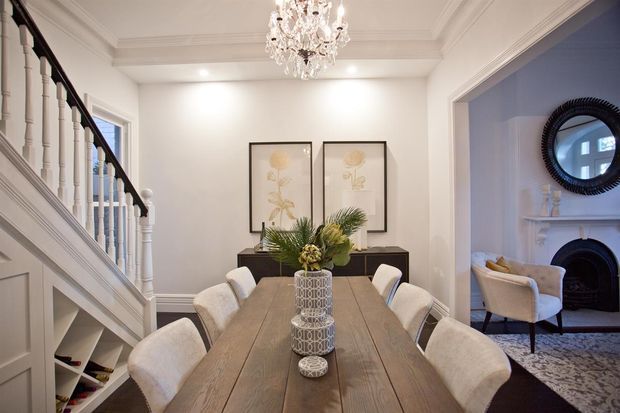 Elegant formal dining room with a long timber dining table and upholstered cream dining chairs, positioned beside a white-painted staircase with turned balusters and paneled wainscoting. The room features white walls and decorative ceiling mouldings, a crystal chandelier, botanical artwork, and a dark console. An adjoining living room with a fireplace and round mirror is visible through a wide opening.