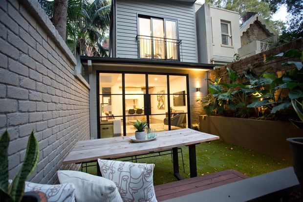 A compact urban courtyard viewed from an outdoor seating area, featuring a two-storey modern extension with dark horizontal cladding, large black-framed glazed doors, and warm interior lighting. The courtyard has a rendered brick boundary wall, raised planter with lush tropical foliage, artificial or closely clipped lawn, and a simple timber outdoor dining table with bench seating.