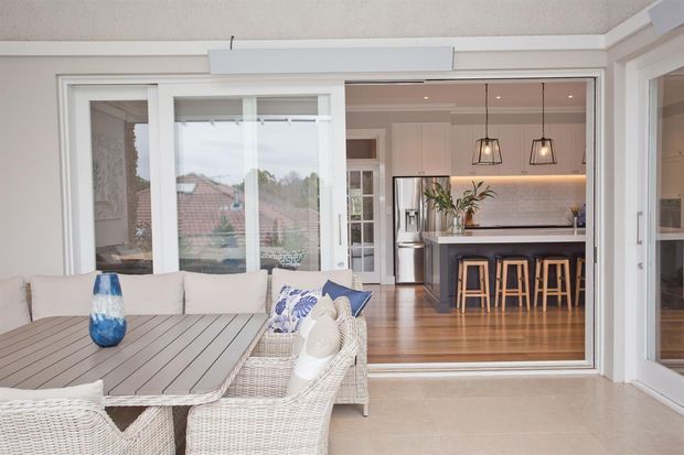 Bright open-plan indoor-outdoor living space viewed from a covered patio or sunroom. A white wicker dining setting occupies the foreground, while large glazed sliding doors open toward a contemporary kitchen with a central island, timber stools, pendant lights, stainless-steel refrigerator, white cabinetry, and timber flooring. The palette is light and neutral with coastal detailing.