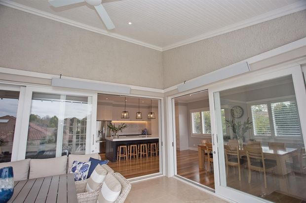 Bright enclosed outdoor living area or sunroom with large white-framed sliding glass doors opening toward a kitchen and dining room. The space has a light tiled floor, woven outdoor seating, a white ceiling fan, textured neutral walls, and a white beadboard-style ceiling. The adjacent kitchen features a dark island with timber stools, while the dining room has timber flooring and a wooden table.