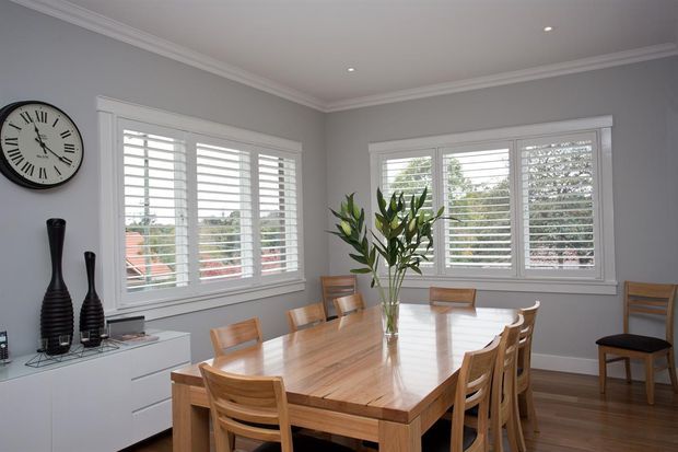 Bright, refined dining room with a large rectangular timber dining table and matching wooden chairs, surrounded by white plantation shutters. Pale grey walls, white ceiling cornices, recessed downlights, timber flooring, and a minimalist white sideboard with black decorative vessels complete the space.