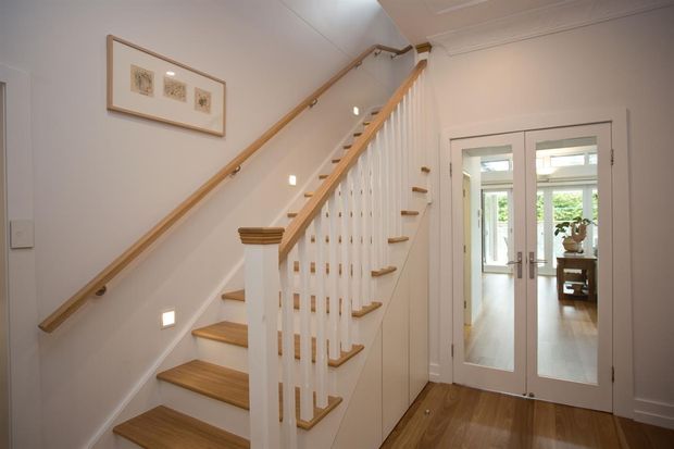 Bright traditional hallway with a timber staircase, white painted balustrade and trim, and warm timber flooring. Double French doors lead toward a light-filled room beyond, while framed artwork and discreet stair lighting add subtle detail.