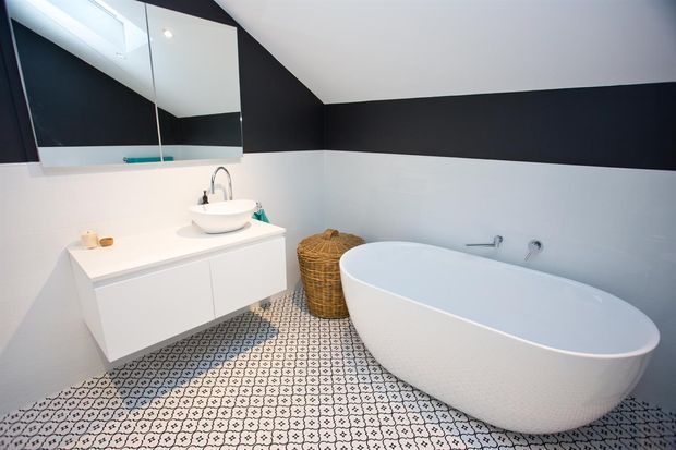 Contemporary bathroom with a freestanding white oval bathtub, floating white vanity with integrated basin, wall-mounted tapware, large mirror, and patterned geometric floor tiles. The room features a bold black horizontal wall band against predominantly white surfaces.