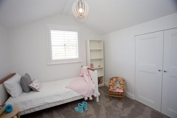 A bright, compact children's bedroom with a single daybed dressed in white bedding and a pink throw, a small bookshelf, a wicker chair holding plush toys, white louvered window shutters, built-in white wardrobe doors, and a decorative pendant light. The room has soft neutral finishes with pink and blue accents and carpeted flooring.