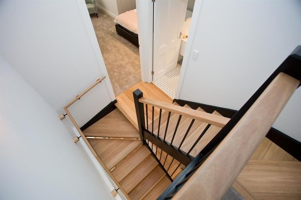 A compact residential stairway viewed from above, with timber stair treads and handrails, black metal balusters and trim, and white walls. The landing opens toward a bedroom visible through a doorway, with tiled flooring glimpsed in an adjacent space.