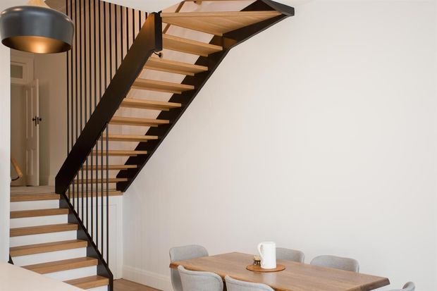 Interior view of a contemporary stairway beside a dining area. The open-riser timber staircase has dark structural stringers and slender black vertical balusters, set against a white wall with a vertical timber-slat feature. A timber dining table and upholstered chairs occupy the foreground, with a modern pendant light overhead.