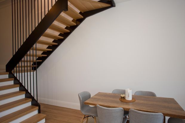 Contemporary open-plan dining area beneath a sculptural timber staircase. The dining table and upholstered chairs sit against a plain light wall, while the staircase features timber treads, dark structural stringers, and slender vertical metal balusters. Timber flooring continues through the space.