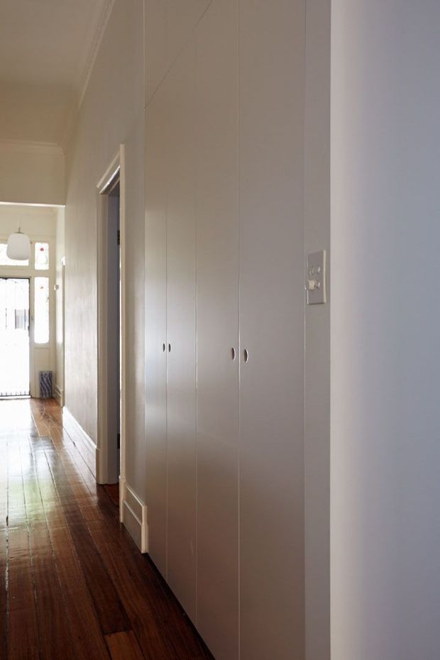A narrow residential hallway with polished dark timber floorboards, pale painted walls, traditional ceiling cornices and a tall bank of built-in white storage cupboards with small dark handles. A glazed entry door and pendant light are visible at the far end, creating a bright, elongated passage.