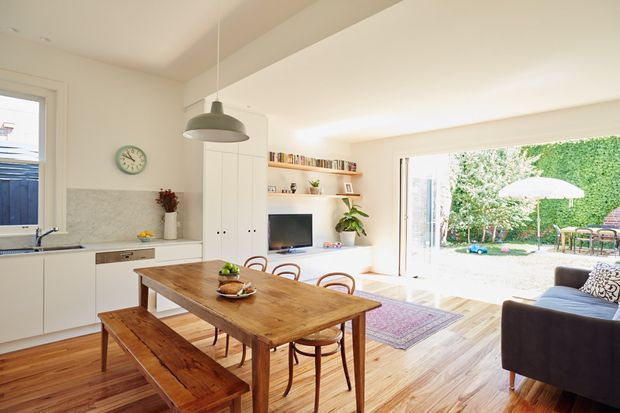 Bright open-plan kitchen and dining area with white cabinetry, a marble-look splashback, warm timber flooring, and a rustic timber dining table with bench seating and bentwood chairs. A compact living area with sofa and wall-mounted TV sits beside large glazed doors opening to a leafy garden and outdoor dining area. The room has a relaxed, light-filled contemporary character with pendant lighting and open shelving.