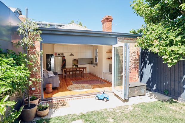 Bright view from a landscaped backyard into a renovated brick dwelling extension, with wide-open glazed doors connecting an outdoor area to a compact open-plan living, dining and kitchen space. The interior features white cabinetry and walls, timber flooring, exposed brick, and a simple pitched roofline, while a dark vertical-board fence and mature planting frame the setting.