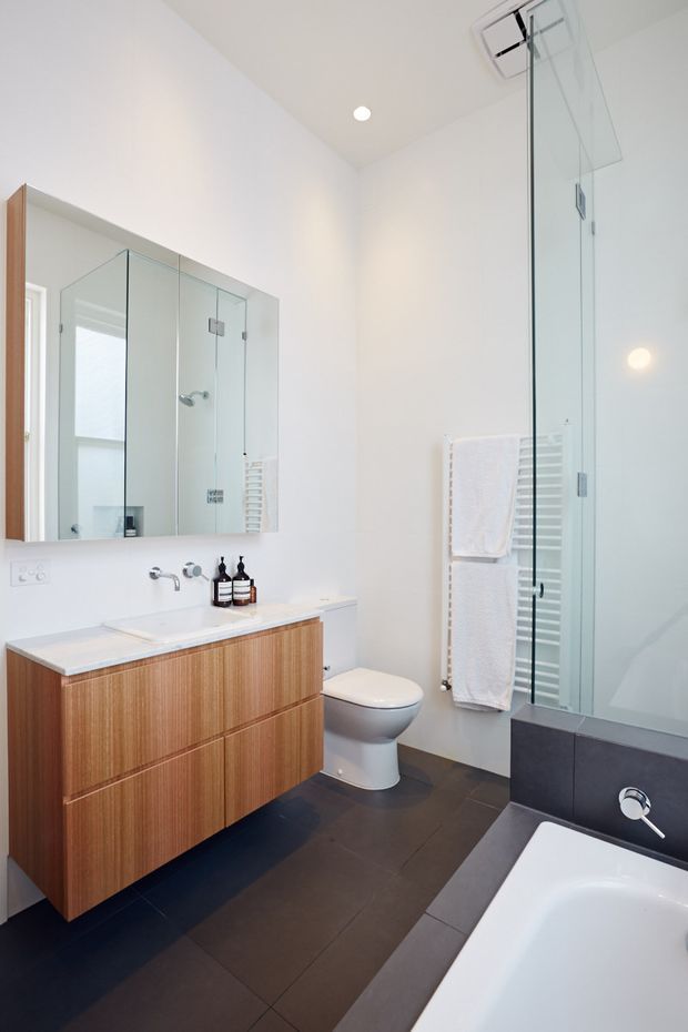 Bright contemporary bathroom with a floating timber vanity, white integrated basin, wall-mounted tapware, mirrored cabinet, toilet, heated towel rail, glass-enclosed shower and freestanding bathtub. Dark large-format floor tiles and dark bath surround contrast with the white walls and warm wood joinery.
