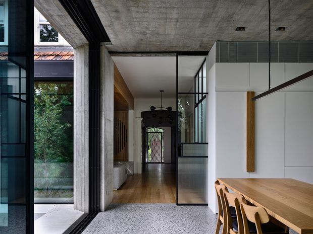 A refined contemporary interior connecting an open dining area with a hallway and glazed courtyard edge. The space features exposed concrete ceilings and structural columns, full-height black-framed glazing, warm timber flooring and wall accents, a large timber dining table with matching chairs, and a darker formal doorway at the end of the corridor. The composition balances raw concrete, glass, timber, and white plaster surfaces.