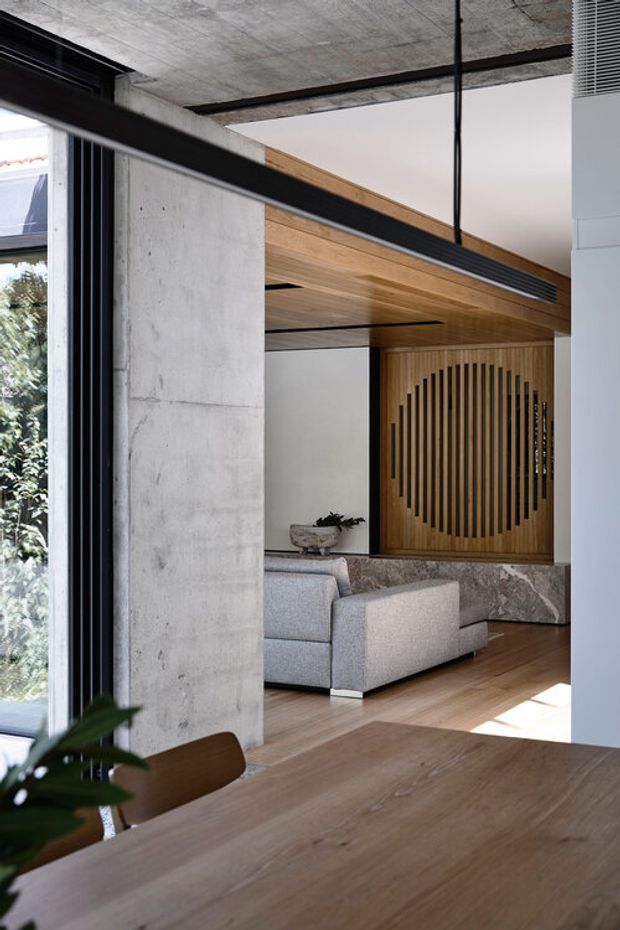 A refined contemporary interior viewed from a dining area, with a large timber dining table in the foreground opening toward a living room. The space features exposed raw concrete walls and ceiling, extensive black-framed glazing, warm timber ceiling and wall panelling, a sculptural slatted timber screen, a stone-clad low wall, and neutral upholstered seating. Strong linear detailing and natural materials create a calm, sophisticated atmosphere.