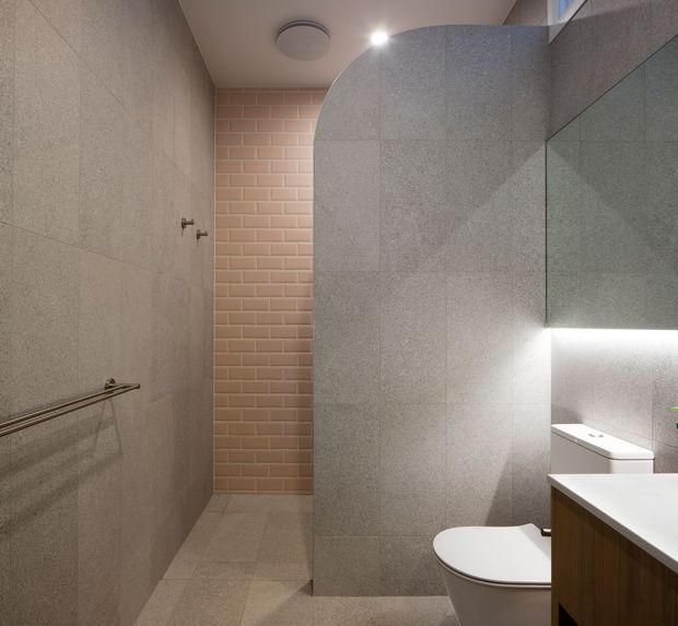 A contemporary bathroom with large grey stone-look wall and floor tiles, a curved tiled partition enclosing the shower area, a vertical strip of soft pink subway tiles, frameless glass, wall-mounted lighting, a toilet, and a timber-veneered vanity with a white benchtop. The space is compact, refined, and softly illuminated.
