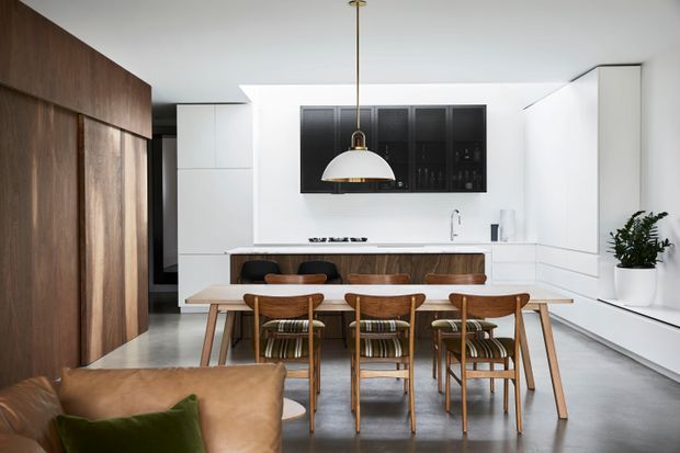 Open-plan contemporary kitchen and dining area with a white minimalist fit-out, dark timber wall panelling and cabinetry, a long timber dining table with matching wooden chairs, and a prominent brass-and-white pendant light. A portion of a living room sofa is visible in the foreground.