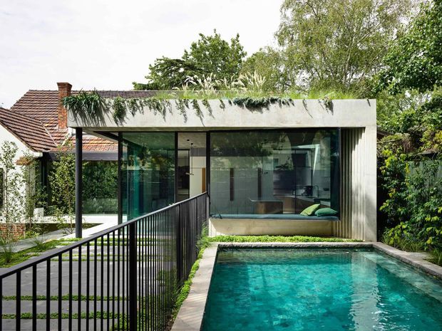 A contemporary glazed pavilion extension beside a swimming pool, connected to an older tiled-roof residence. The modern structure has a flat concrete roof covered with trailing greenery, large floor-to-ceiling windows, dark slender framing, and a covered outdoor walkway with black metal balustrades. Lush garden planting surrounds the pool and building, creating a strong contrast between the heritage-style house and the crisp modern addition.