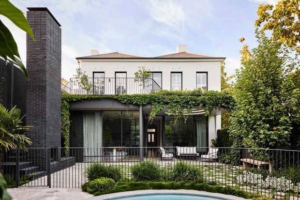 Exterior view of a refined two-storey residence with a pale rendered upper level, low hipped roof, extensive black-framed glazing, and a dark brick vertical chimney or stair-tower element. The ground floor opens onto a landscaped terrace and pool, with black metal balustrades, lush greenery, climbing vines, and a restrained contemporary composition.