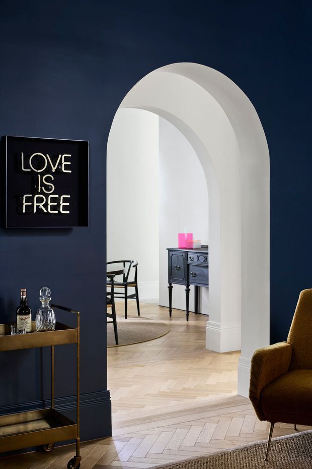 A moody, refined interior view framed by a large white arched opening. The foreground living area has deep navy walls, pale herringbone timber flooring, a mustard upholstered armchair, a brass-toned drinks trolley, and a framed neon-style sign. Through the arch is a light dining area with black chairs and a dark vintage-style side cabinet. White architectural mouldings and the curved arch contrast strongly with the dark wall.
