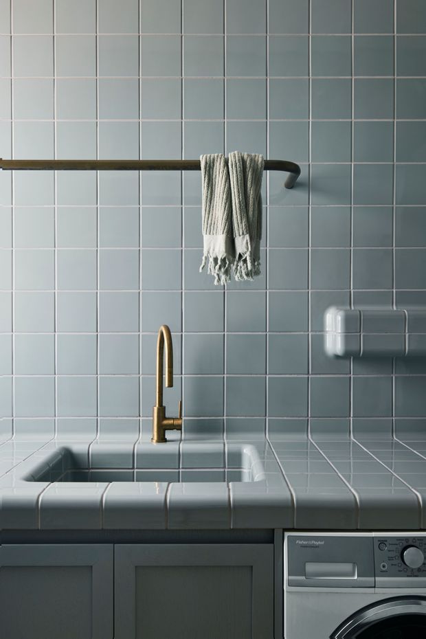 Close-up of a compact laundry room with a deep tiled utility sink, grey-blue square wall and benchtop tiles, dark cabinetry below, a brass gooseneck tap, wall-mounted towel rail, and a front-loading washing machine. The image has a refined, moody, utilitarian interior aesthetic.