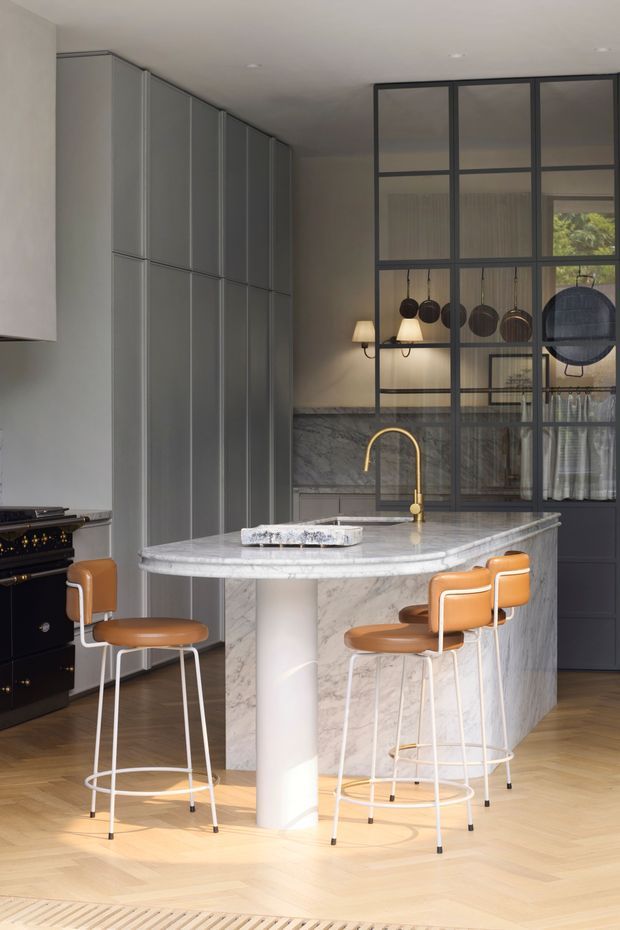 A refined contemporary kitchen featuring a large veined marble island with a rounded edge and cylindrical support, four caramel leather bar stools, tall matte grey integrated cabinetry, and a dark freestanding range. A black-framed glazed partition reveals hanging cookware and a softly lit adjoining space. Pale timber parquet flooring, warm brass tapware, and restrained recessed lighting create an elegant, warm interior.
