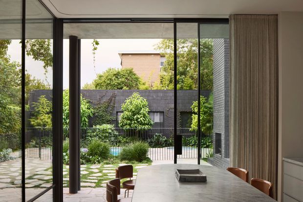 A contemporary dining area viewed from inside, with a long stone dining table and leather chairs positioned beside full-height black-framed glazing. The glazing opens visually onto a landscaped courtyard with stepping-stone paving, dense planting, a pool glimpse, and dark masonry boundary walls. A textured vertical wall finish and integrated cabinetry are visible on the right.