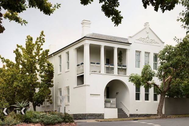 Street-facing view of a substantial white two-storey residence with a formal symmetrical facade, steep gabled section, slate-like hipped roof, tall sash-style windows, classical columns, upper-level balcony, arched entry passage, decorative gable relief, and dark stone base. Mature trees and restrained front landscaping frame the building.