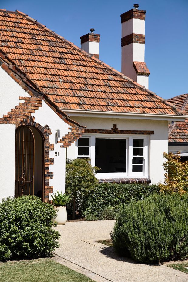 Exterior view of a single-storey white-rendered house with a steep terracotta tiled pitched roof, prominent brick chimneys, decorative brickwork around the arched entry and windows, and a landscaped front garden with a paved path. The facade has a traditional bungalow character and strong Mediterranean-influenced detailing.