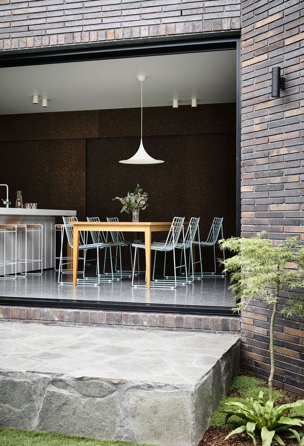 A contemporary dining area viewed from an outdoor courtyard through a large open glazed opening. The interior features a timber dining table surrounded by white wire-frame chairs, a dark textured feature wall, a white pendant light, polished grey flooring, and a compact kitchen counter at left. The surrounding exterior is clad in dark grey-brown brick, with a raised natural-stone terrace and planted garden elements in the foreground.