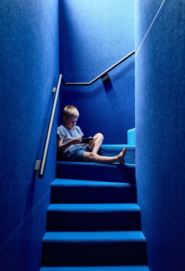 A narrow, enclosed stairway clad almost entirely in vivid blue carpet and blue wall finishes. Metal handrails run along both sides and continue up the stairwell, with a small child seated on the upper steps using a handheld device. The image has a dramatic, monochromatic blue appearance and strong vertical perspective.