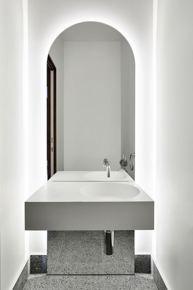 A narrow, minimalist bathroom with a floating white vanity featuring an integrated basin and chrome tapware. A large rounded-arch mirror spans the wall above the vanity, while grey terrazzo tiles cover the floor and lower wall area. White walls, concealed plumbing, and recessed lighting create a clean, highly pared-back composition.