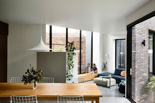 Bright contemporary open-plan dining and living area with a large solid timber dining table, sculptural curved plaster rangehood or partition element, black-framed floor-to-ceiling glazing, and an adjoining lounge with a dark blue sofa. Exposed dark brickwork is visible through the open glazed wall, with indoor plants adding greenery. The space has a clean, airy, modern feel and strong indoor-outdoor visual connection.
